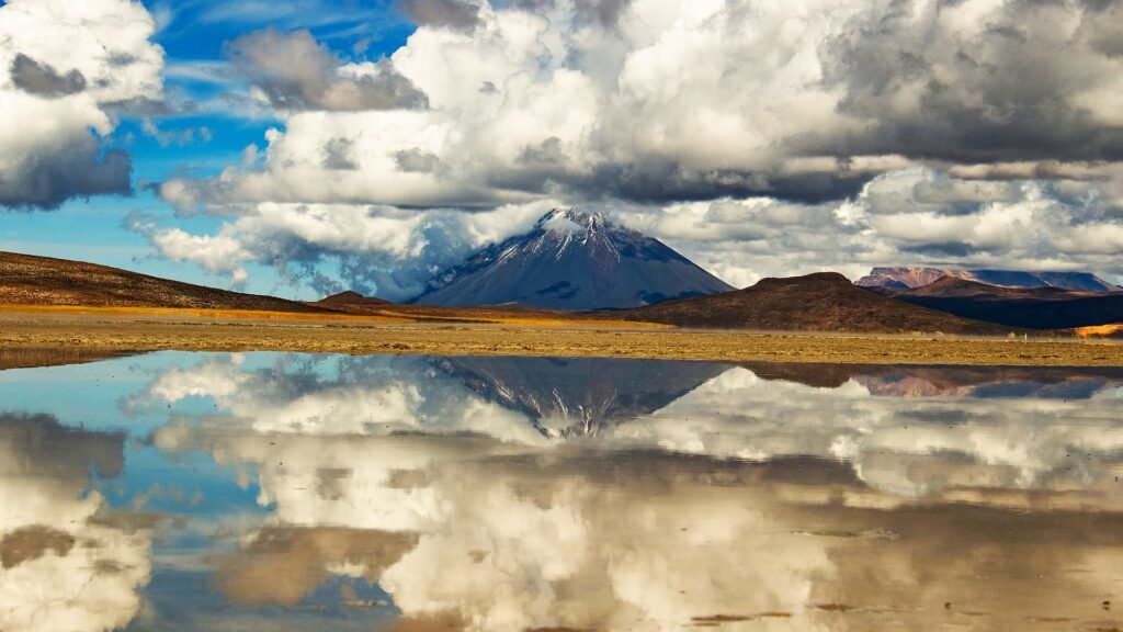 Laguna cristalina que actúa como un espejo perfecto reflejando nubes blancas y un volcán nevado al fondo